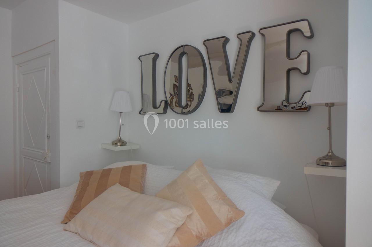 Lit avec coussins beige clair, encadré par deux lampes de chevet, et décoration murale formant le mot ’LOVE’.