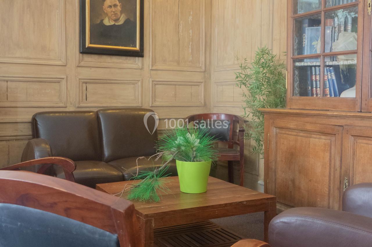 Salon avec fauteuils en cuir, table basse en bois, plante verte, bibliothèque et tableau sur un mur lambrissé.