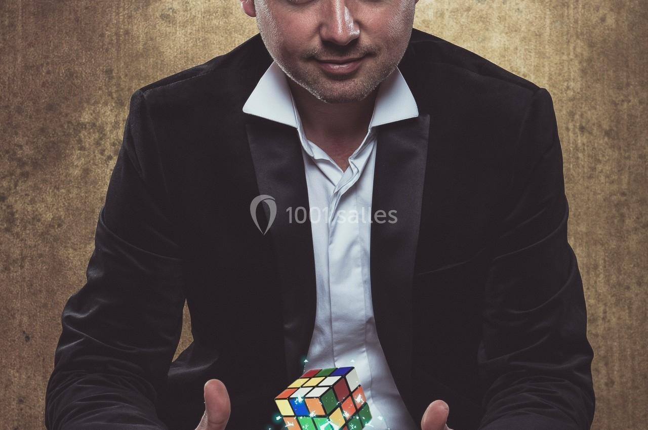Un homme en costume noir tient un Rubik's Cube lumineux entre ses mains devant un fond texturé.