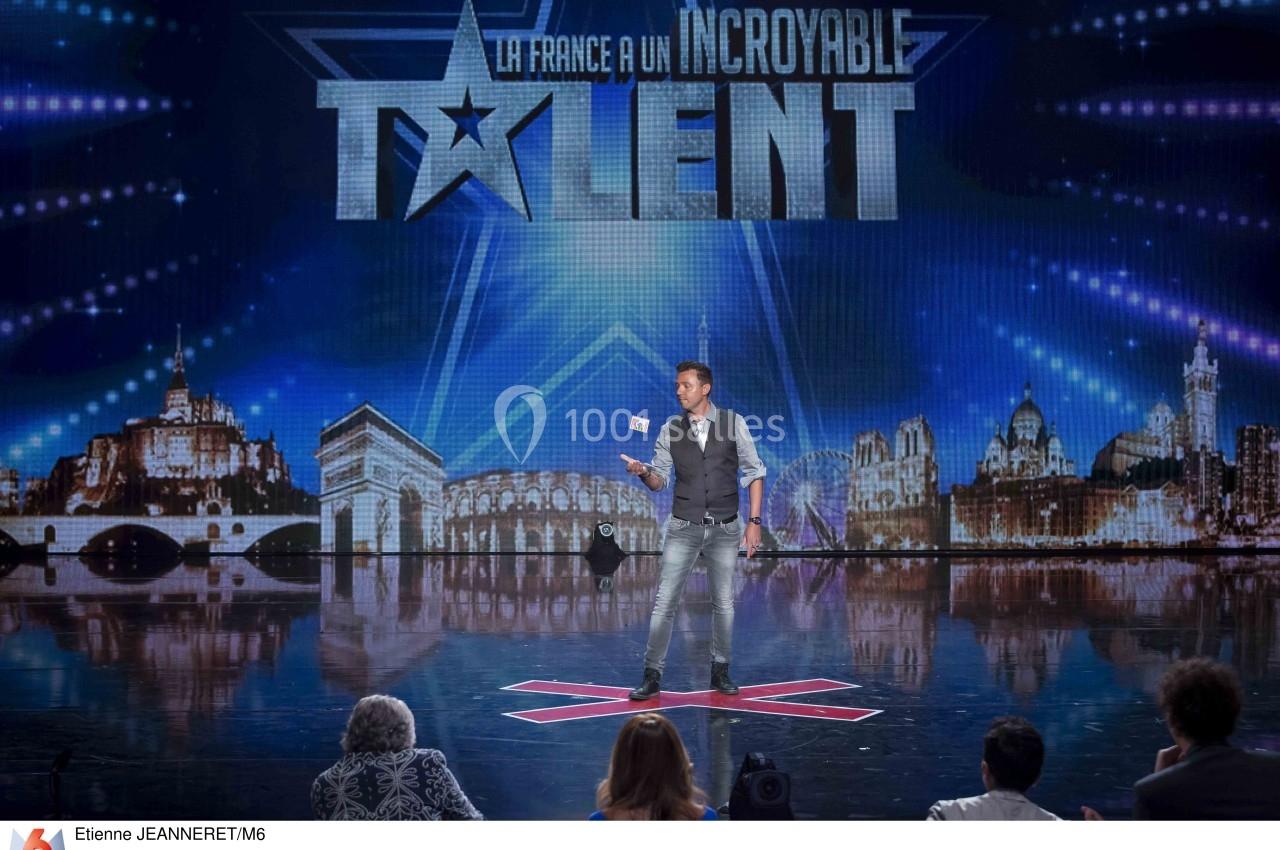 Un homme réalise un numéro de magie sur une scène illuminée, devant un jury et un décor urbain en arrière-plan.