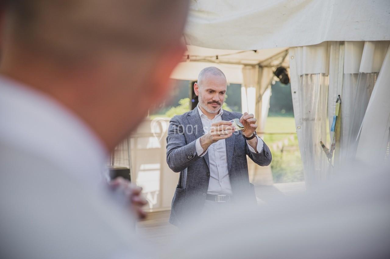 Un homme en costume gris effectue un tour de magie avec une carte dans un espace extérieur sous une tente.