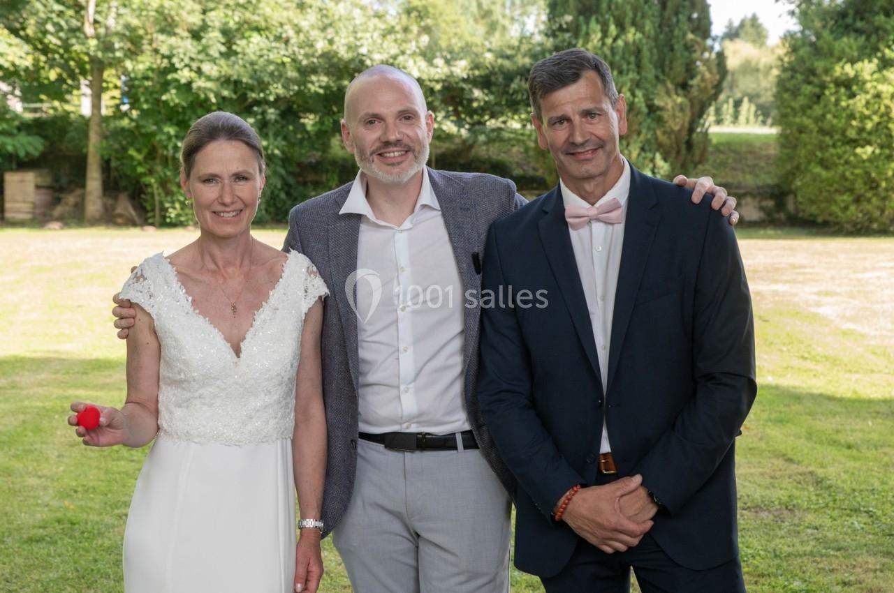 Une femme en robe blanche et deux hommes en costume posent dans un jardin verdoyant par temps clair.