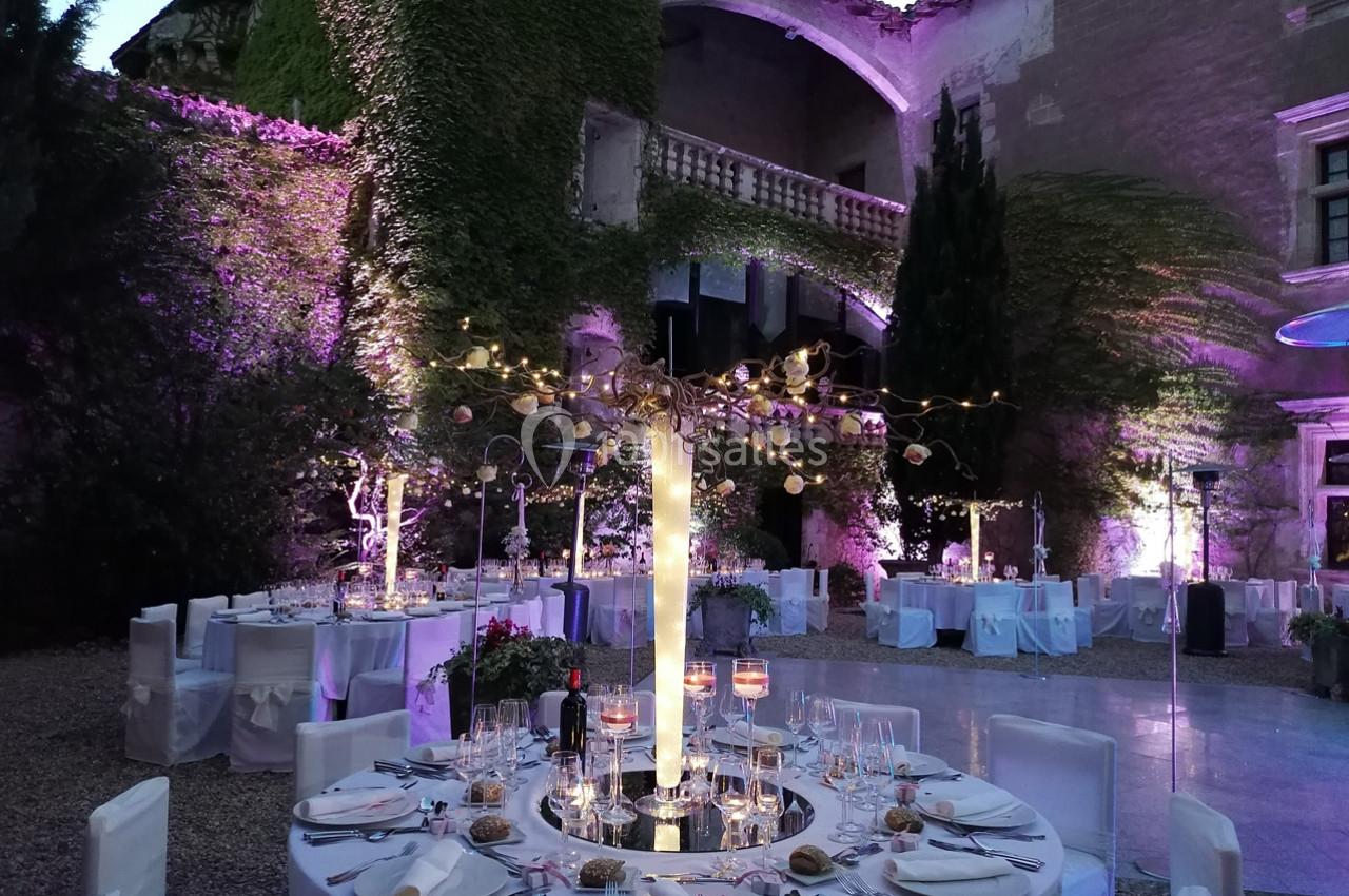 Tables dressées pour un dîner en plein air dans une cour illuminée, entourée de murs couverts de végétation.