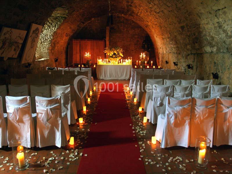 Salle voûtée décorée pour une cérémonie, avec chaises blanches, tapis rouge, bougies et pétales au sol.
