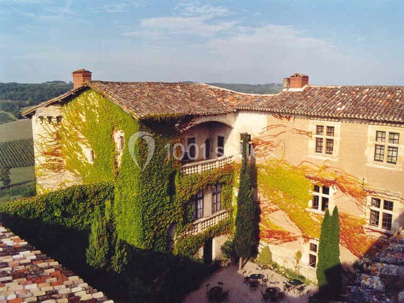 Façade d'un bâtiment ancien couvert de vigne vierge, avec une cour et un paysage vallonné en arrière-plan.