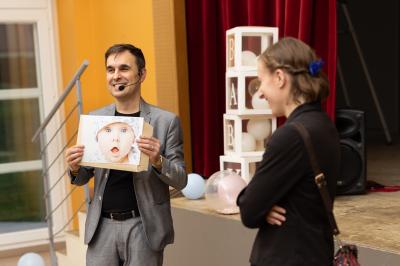 Miniature Hike - Magicien Close Up Et Mentaliste #2 Un homme tenant une photo d'un bébé s'adresse à une femme dans une salle avec rideaux rouges et décorations.