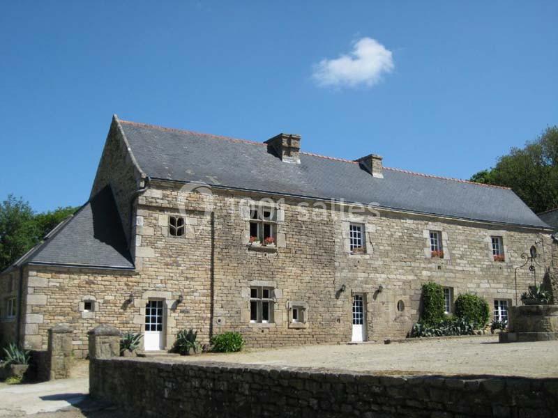 Maison en pierre traditionnelle avec toit en ardoise, entourée de végétation et d'une cour pavée.
