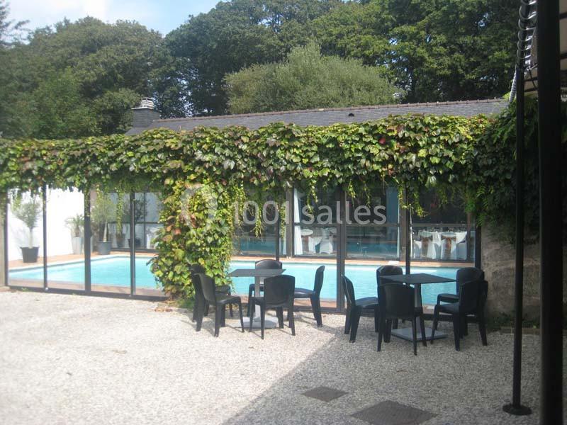 Piscine intérieure visible à travers une baie vitrée entourée de murs recouverts de plantes grimpantes, avec des tables…