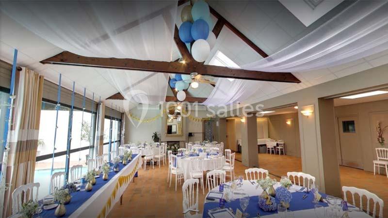 Salle de réception décorée avec des tables dressées, des guirlandes et des ballons bleus et blancs suspendus au plafond.