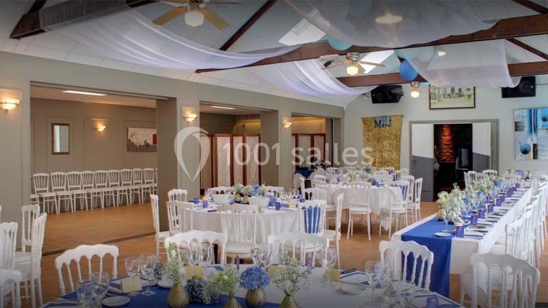 Salle de réception décorée avec des tables dressées, nappes blanches, fleurs bleues et voilages suspendus au plafond.