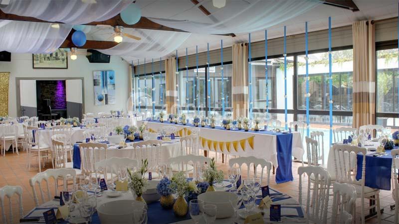 Salle décorée pour un événement, avec tables dressées, nappes bleues et blanches, et guirlandes suspendues.