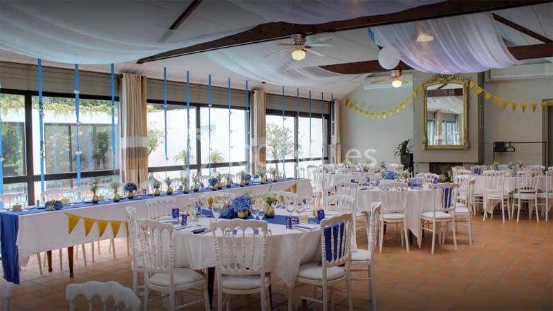Salle décorée pour un événement avec tables dressées, nappes blanches, guirlandes jaunes et bleues, et plafond drapé.