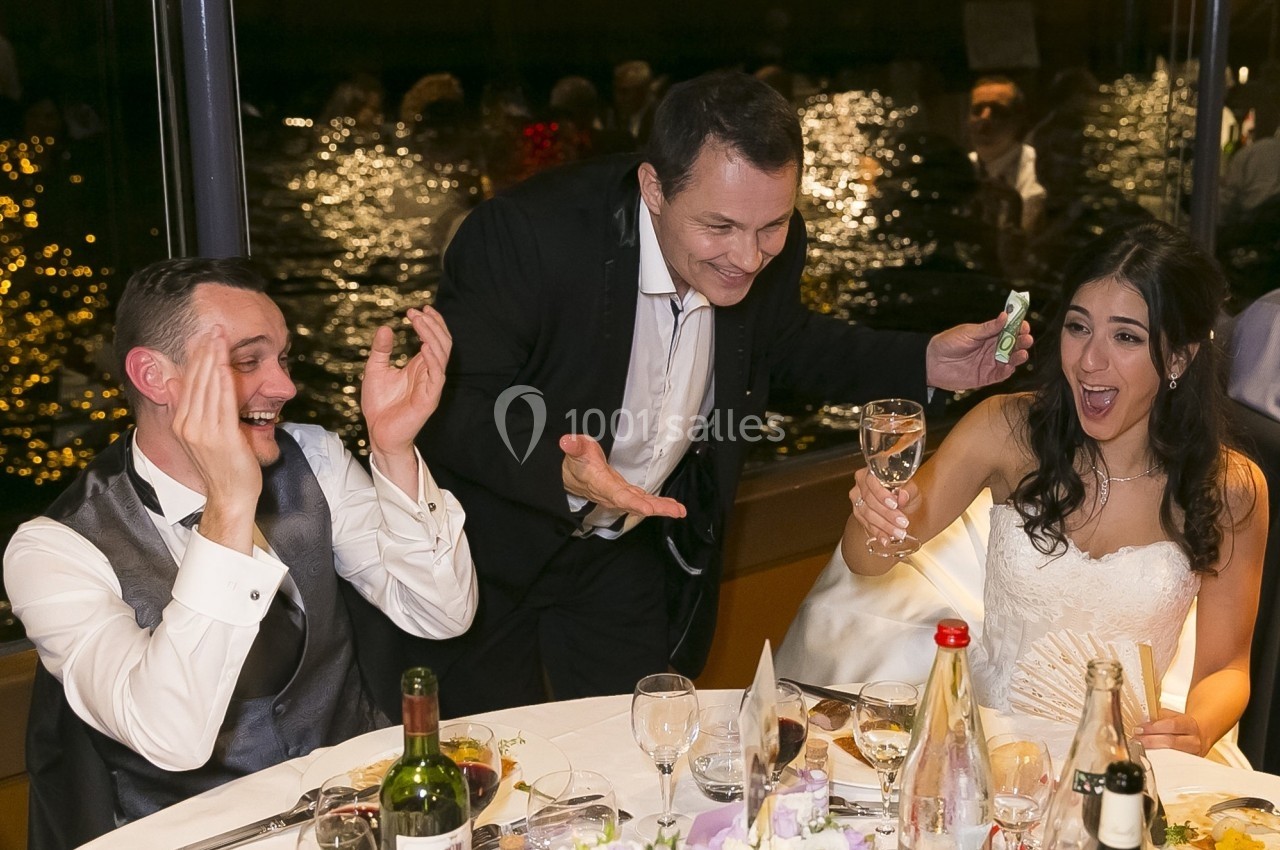 Un couple de mariés assis rit avec un invité debout qui tient un verre et un billet, lors d'une réception festive.
