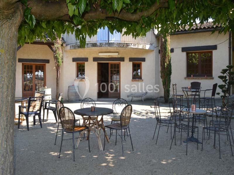 Terrasse ombragée avec des tables et des chaises en métal devant un bâtiment à façade beige et volets en bois.