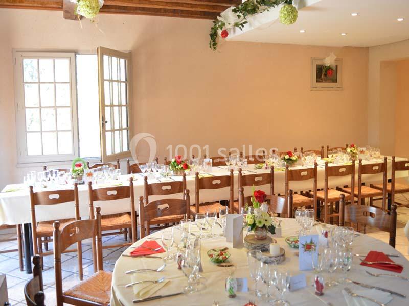 Salle lumineuse avec tables dressées pour un repas, décorée de fleurs et de nappes blanches, chaises en bois autour.