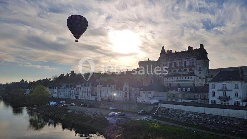 Montgolfière survolant un château et des maisons au bord d'une rivière sous un ciel partiellement nuageux.