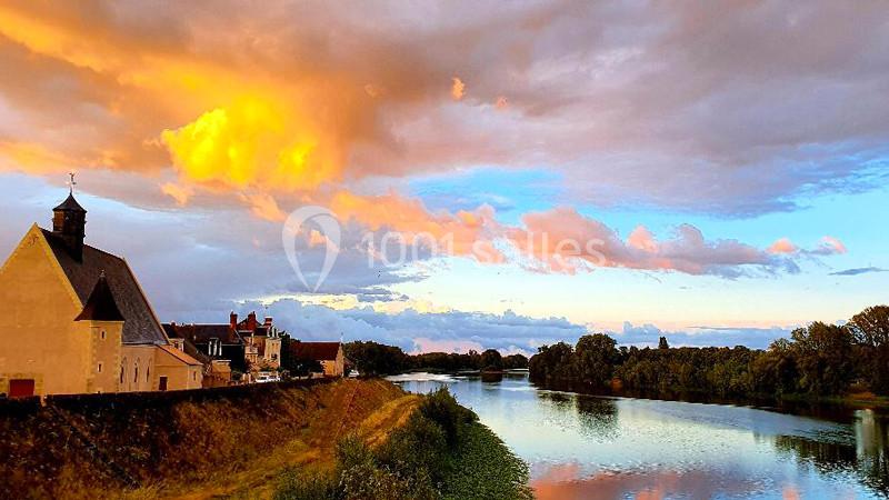 Vue d'un village au bord d'une rivière au coucher du soleil, avec des nuages colorés et un ciel partiellement dégagé.