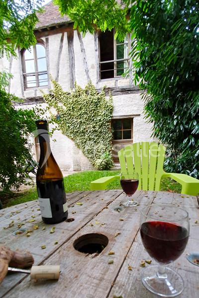 Table en bois avec deux verres de vin rouge, une bouteille ouverte, dans un jardin devant une maison à colombages.