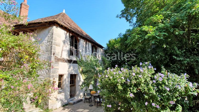 Façade d'une maison en pierre avec toit en tuiles, entourée de végétation et d'arbustes fleuris sous un ciel bleu.