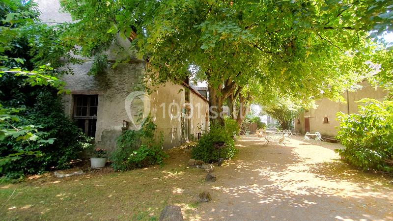 Cour ombragée avec arbres, bâtiments en pierre et mobilier de jardin blanc disposé sur un sol en terre battue.