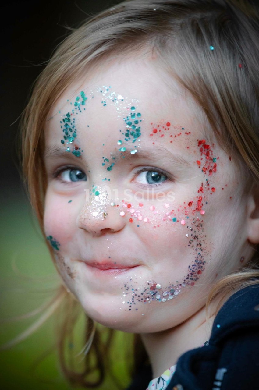 Fillette au visage maquillé de paillettes turquoise, rouges et argentées, regardant vers la droite sur un fond flou vert et sombre.