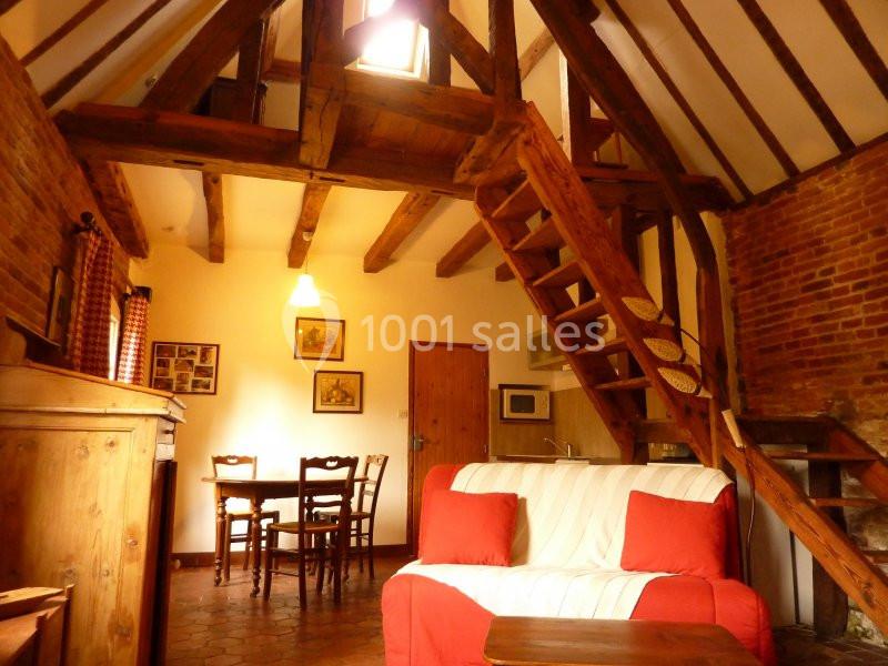 Salon rustique avec poutres apparentes, escalier en bois, canapé rouge et blanc, table à manger et kitchenette.