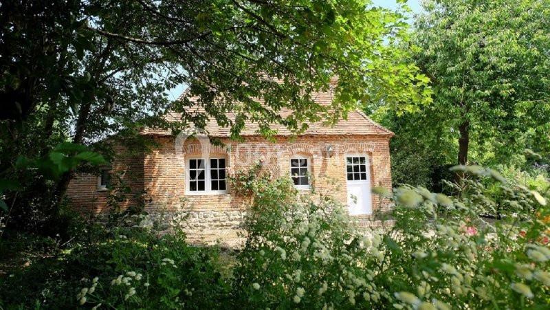 Maison en briques rouges avec toit en tuiles, entourée de verdure et d'arbres sous un ciel ensoleillé.