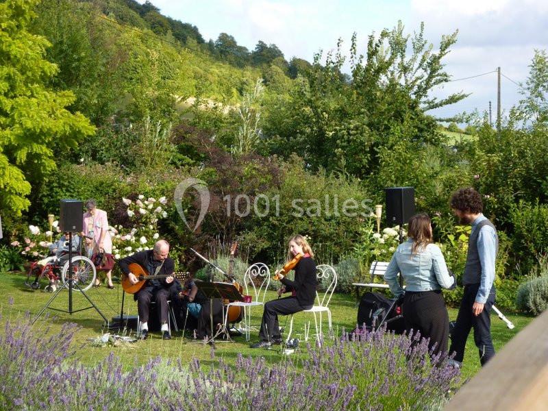 Un groupe de musiciens joue dans un jardin verdoyant, entouré de fleurs et d'arbustes, sous un ciel partiellement nuageux.