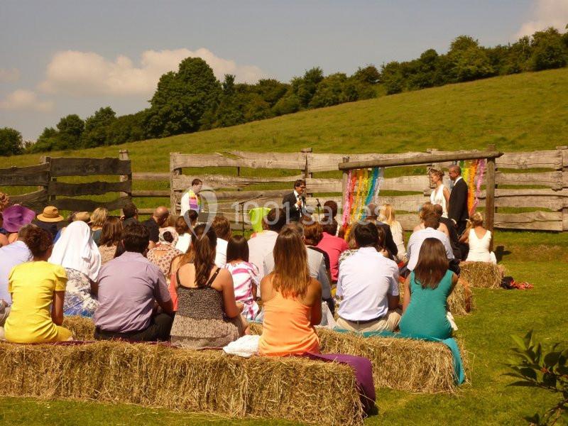 Un mariage en plein air avec des invités assis sur des bottes de foin face à un officiant et un couple devant une arche…