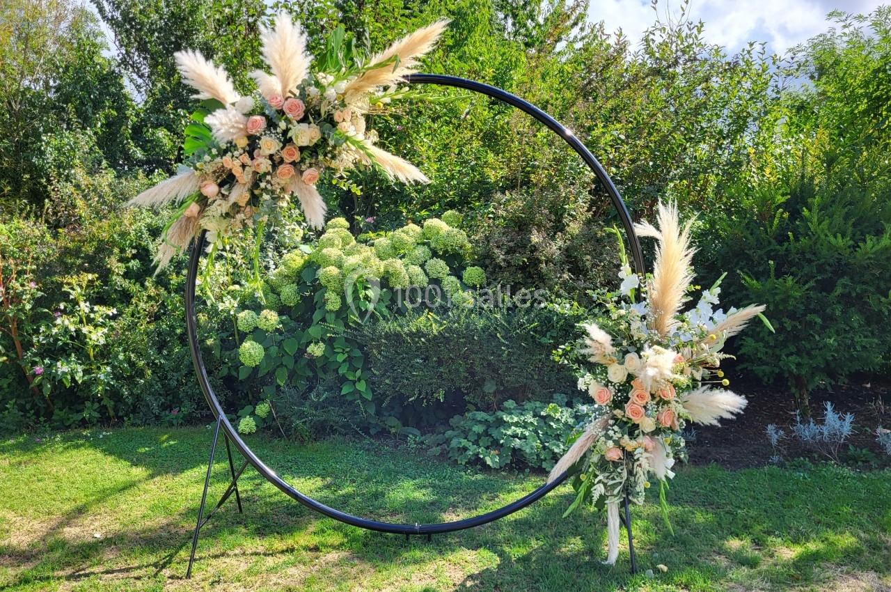 Cercle métallique décoré de fleurs et de pampas, installé dans un jardin verdoyant sous un ciel partiellement nuageux.