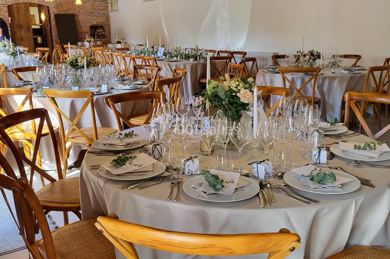 Salle de réception décorée avec des tables rondes dressées, chaises en bois et éclairage chaleureux.