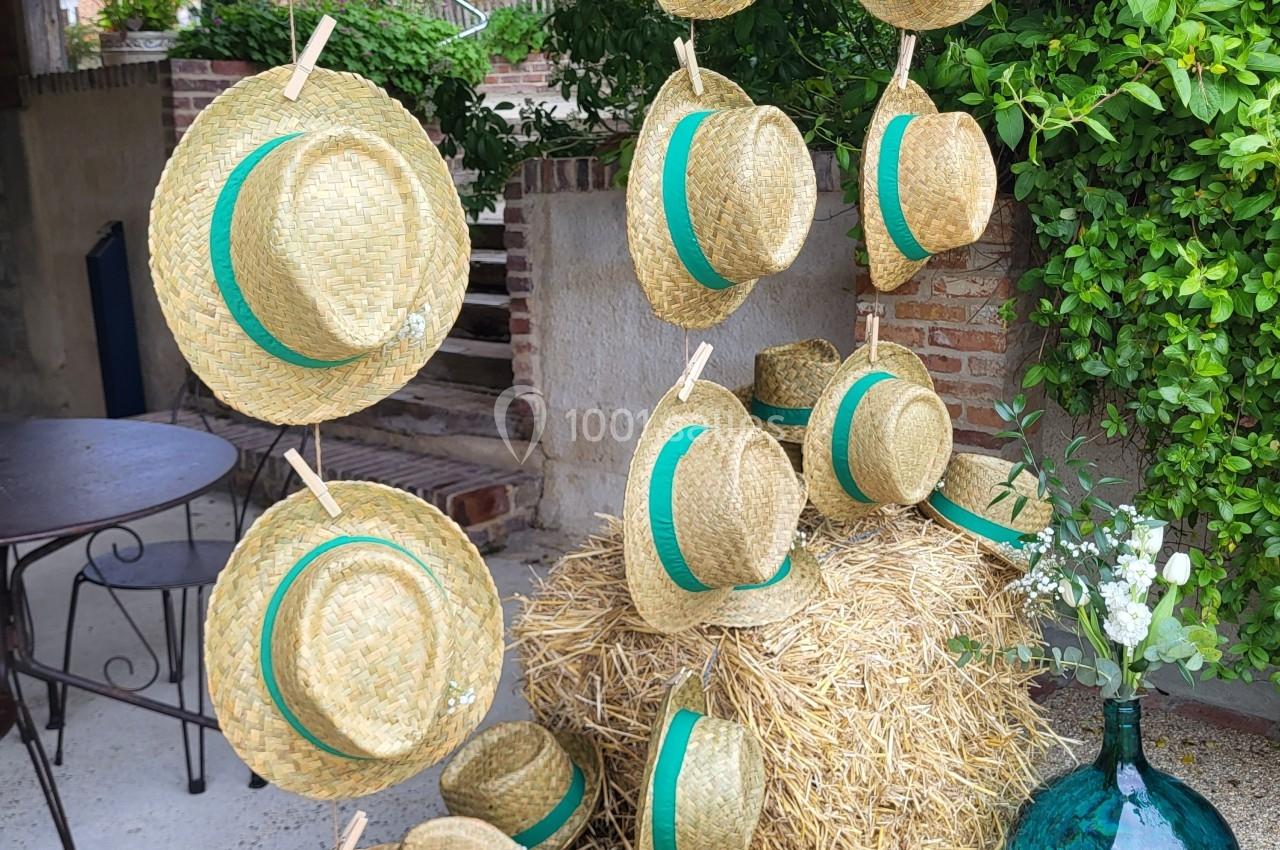 Chapeaux de paille avec rubans verts suspendus et posés sur des bottes de foin, près d'une jarre en verre turquoise.