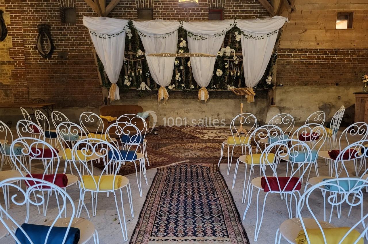 Salle de réception décorée pour un événement, avec des chaises blanches alignées et une arche ornée de fleurs et de drapés.