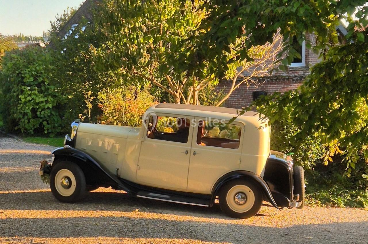Voiture ancienne beige garée sur une allée gravillonnée, entourée de verdure et d'arbres sous une lumière douce.