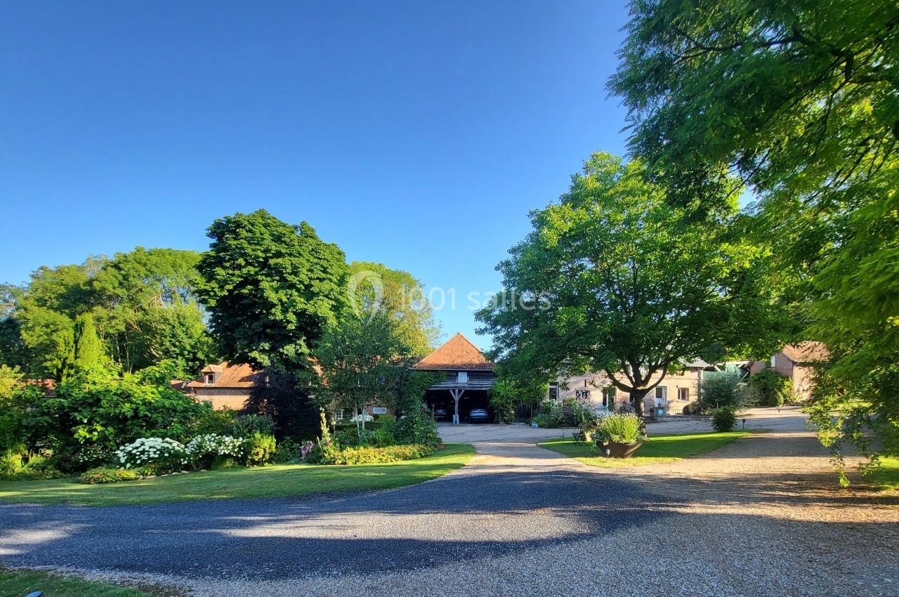 Cour ensoleillée avec allée gravillonnée, arbres verdoyants et bâtiments en pierre sous un ciel bleu clair.