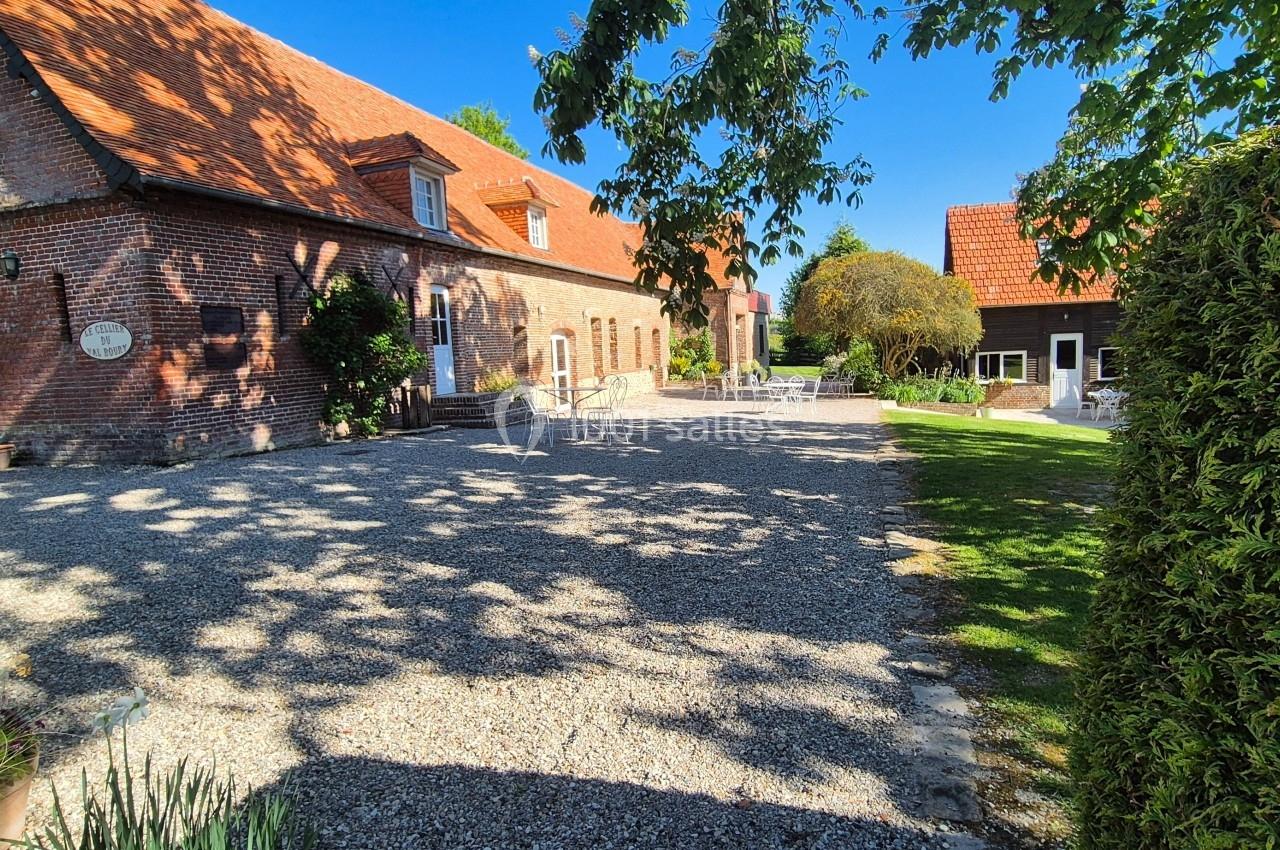 Cour gravillonnée bordée de bâtiments en briques rouges et de verdure, sous un ciel bleu dégagé.