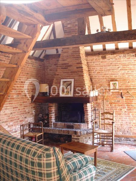 Salon rustique avec murs en briques, cheminée centrale, poutres apparentes, canapé à carreaux et mobilier en bois.