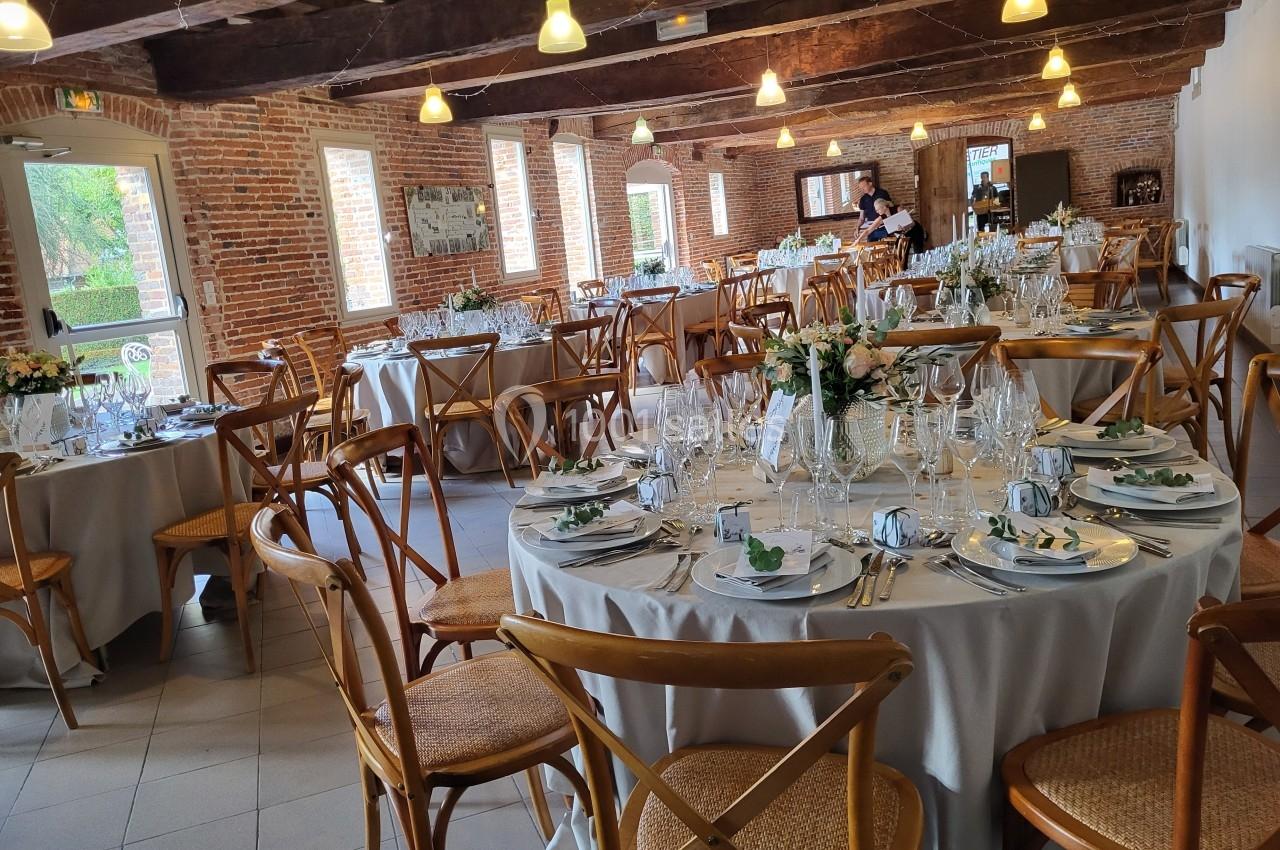 Salle de réception avec tables rondes dressées, nappes blanches, chaises en bois et décoration florale.