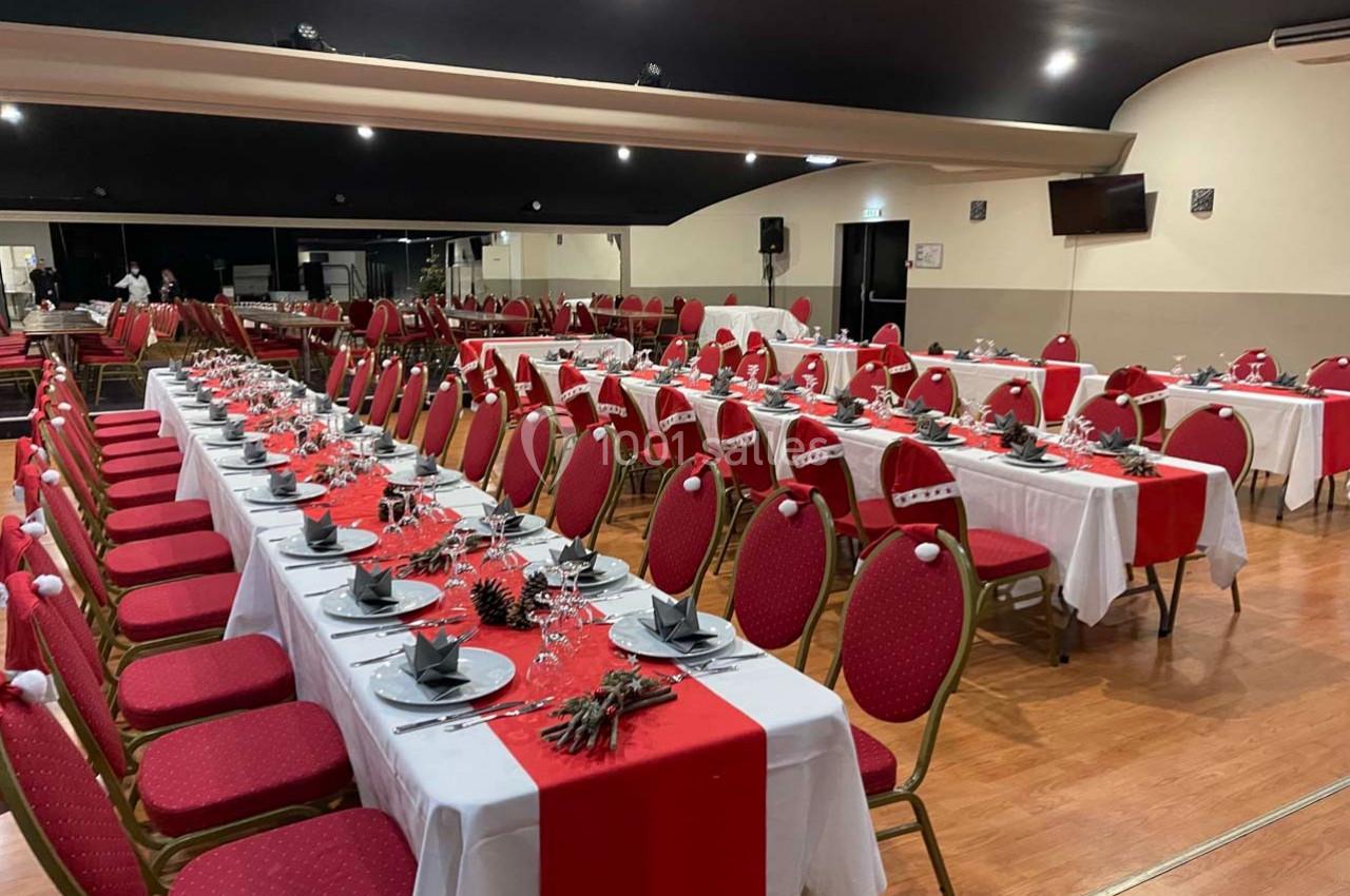 Salle de réception décorée avec des tables alignées, nappes blanches, chemins de table rouges et chaises assorties.