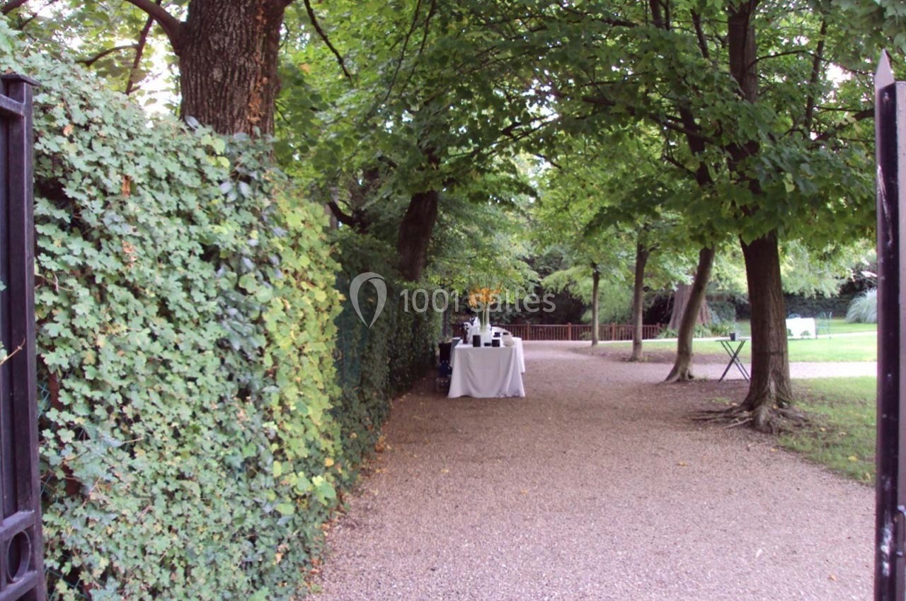 Allée bordée d'arbres et de haies, avec une table dressée sous les arbres au centre de l'image.