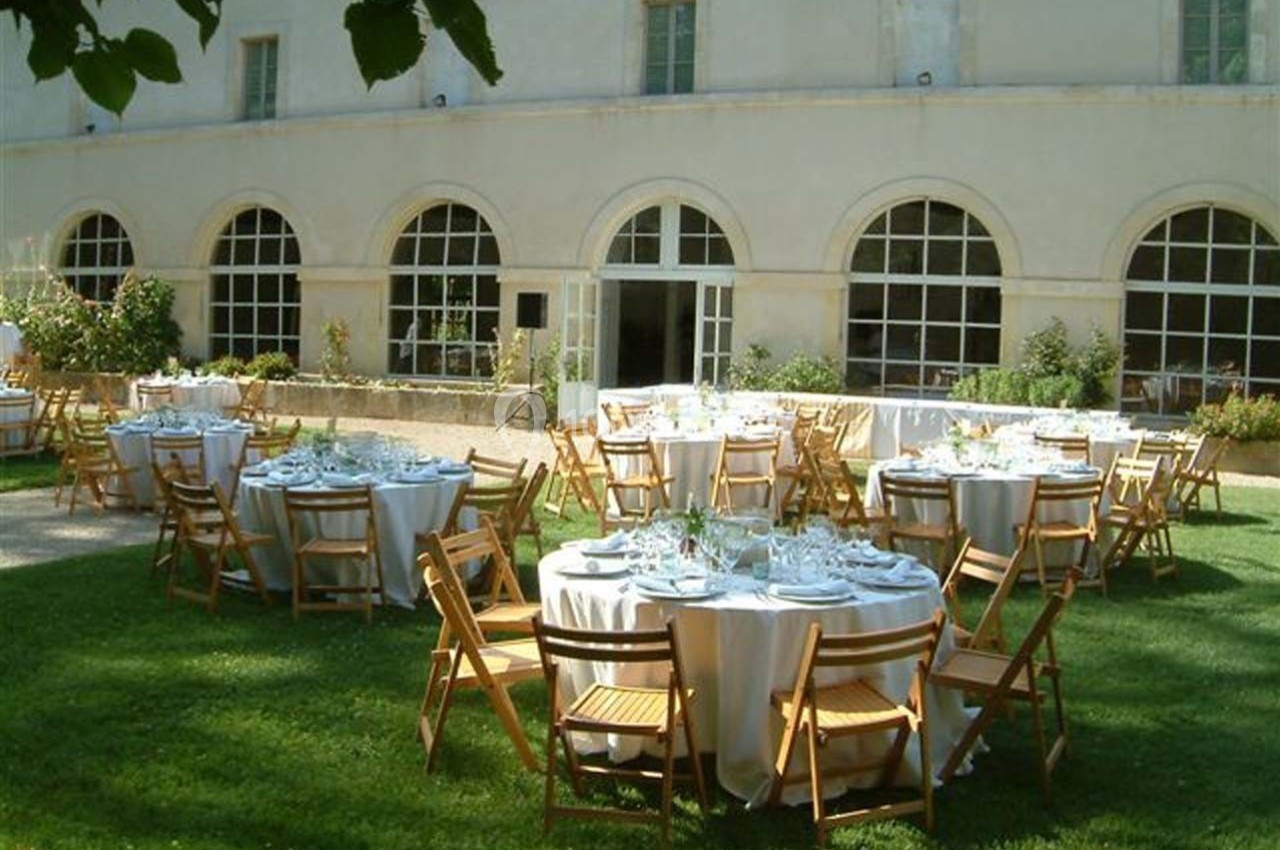 Tables rondes dressées avec nappes blanches et chaises en bois disposées sur une pelouse devant un bâtiment ancien.
