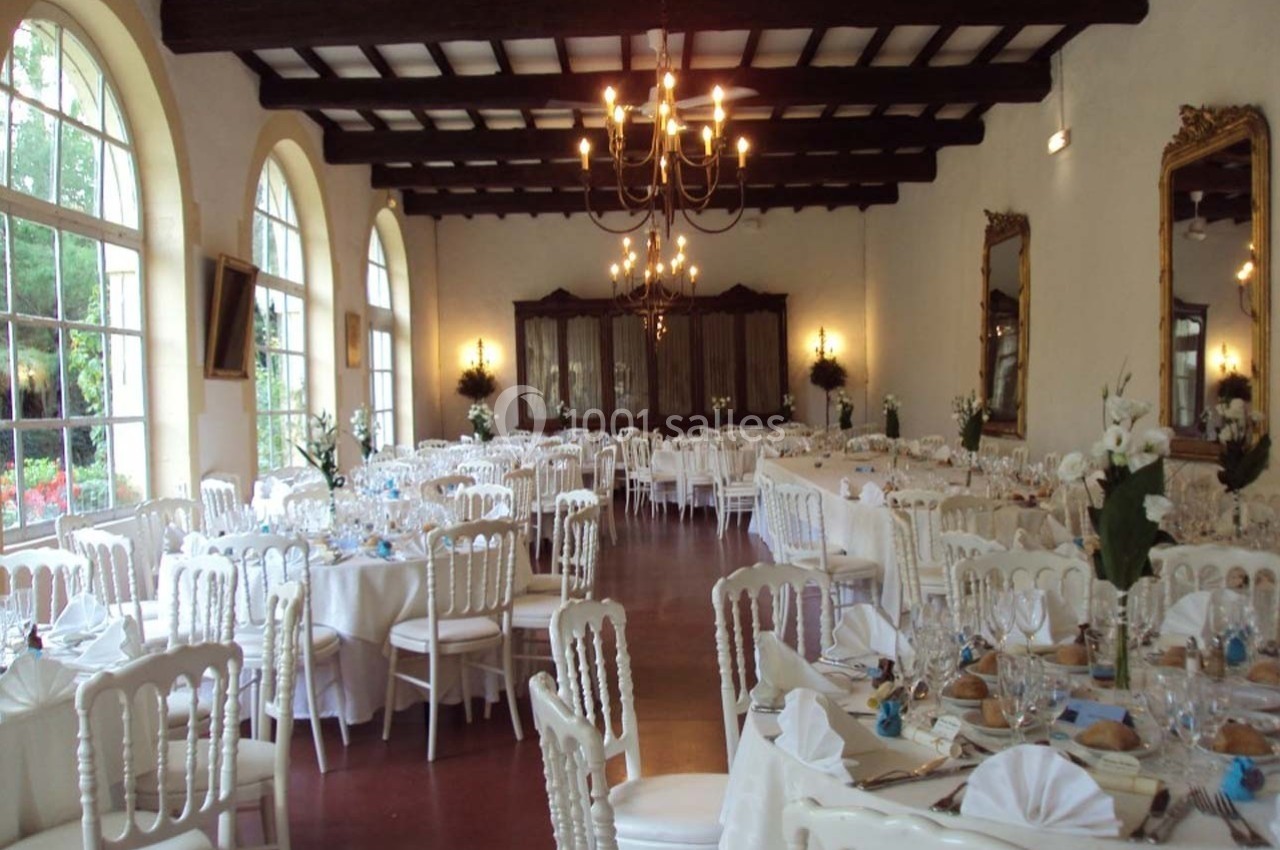 Salle de réception élégante avec tables rondes dressées, chaises blanches et grandes fenêtres laissant entrer la lumière.