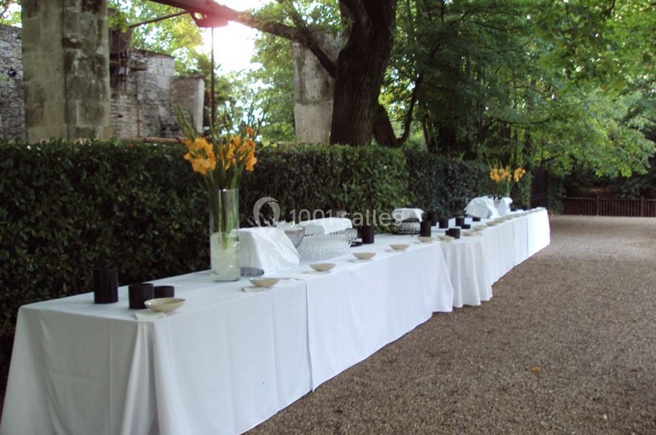 Table dressée en extérieur avec nappes blanches, vaisselle et vases de fleurs jaunes, entourée de verdure.