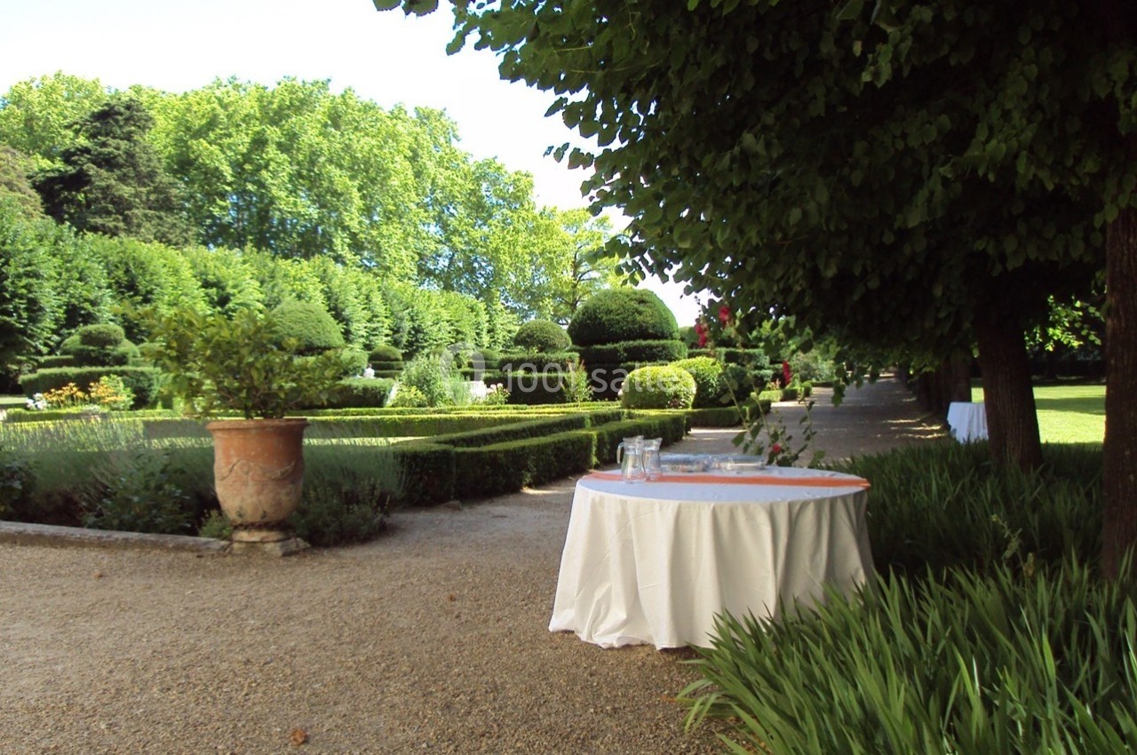 Table ronde avec nappe blanche dans un jardin verdoyant, entourée d'arbres, de haies taillées et d'une allée gravillonnée.