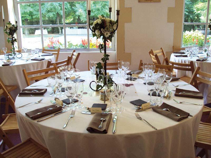 Table ronde élégamment dressée avec nappes blanches, couverts, verres et centre de table floral dans une salle lumineuse.
