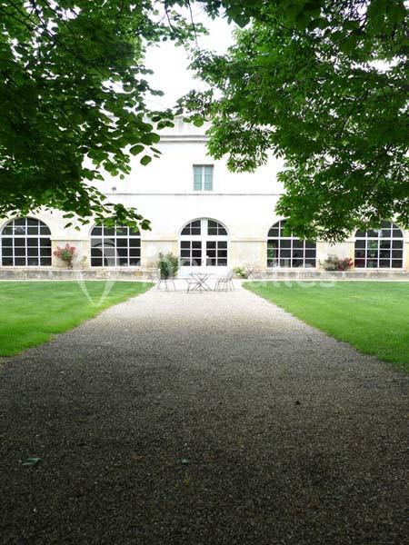 Allée de gravier menant à un bâtiment blanc avec des arches vitrées, entouré de pelouses et d'arbres.