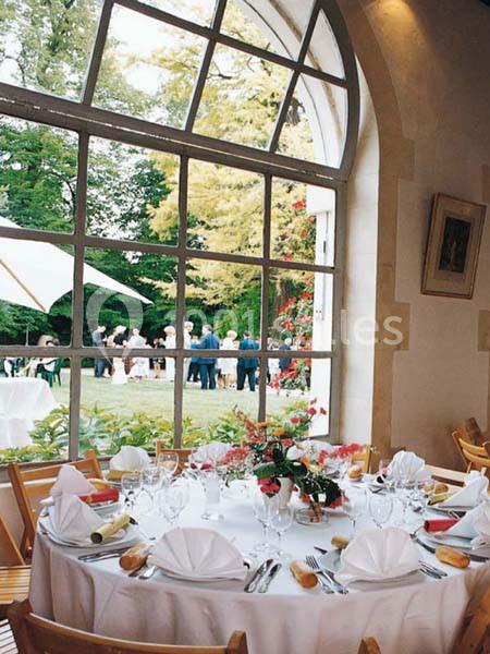 Table dressée avec nappes blanches et vaisselle élégante, vue sur un jardin et des invités à travers une grande baie vitrée.