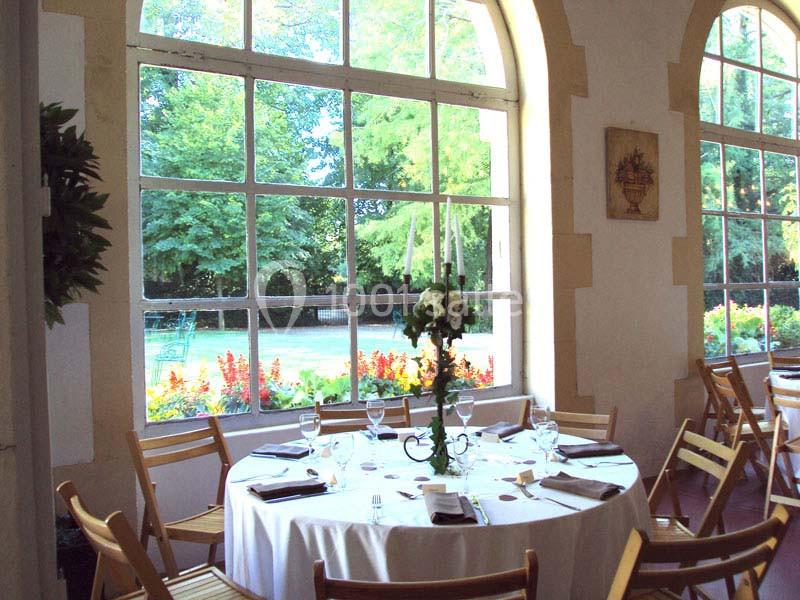 Salle lumineuse avec une table ronde dressée, entourée de chaises en bois, donnant sur un jardin verdoyant par de grandes…