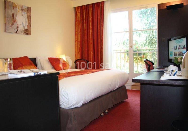 Chambre d'hôtel avec lit double, bureau, télévision, rideaux rouges et grande fenêtre donnant sur un espace verdoyant.