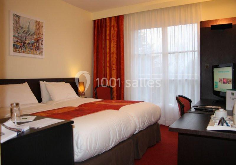 Chambre d'hôtel avec lit double, bureau, télévision, rideaux rouges et grande fenêtre laissant entrer la lumière naturelle.